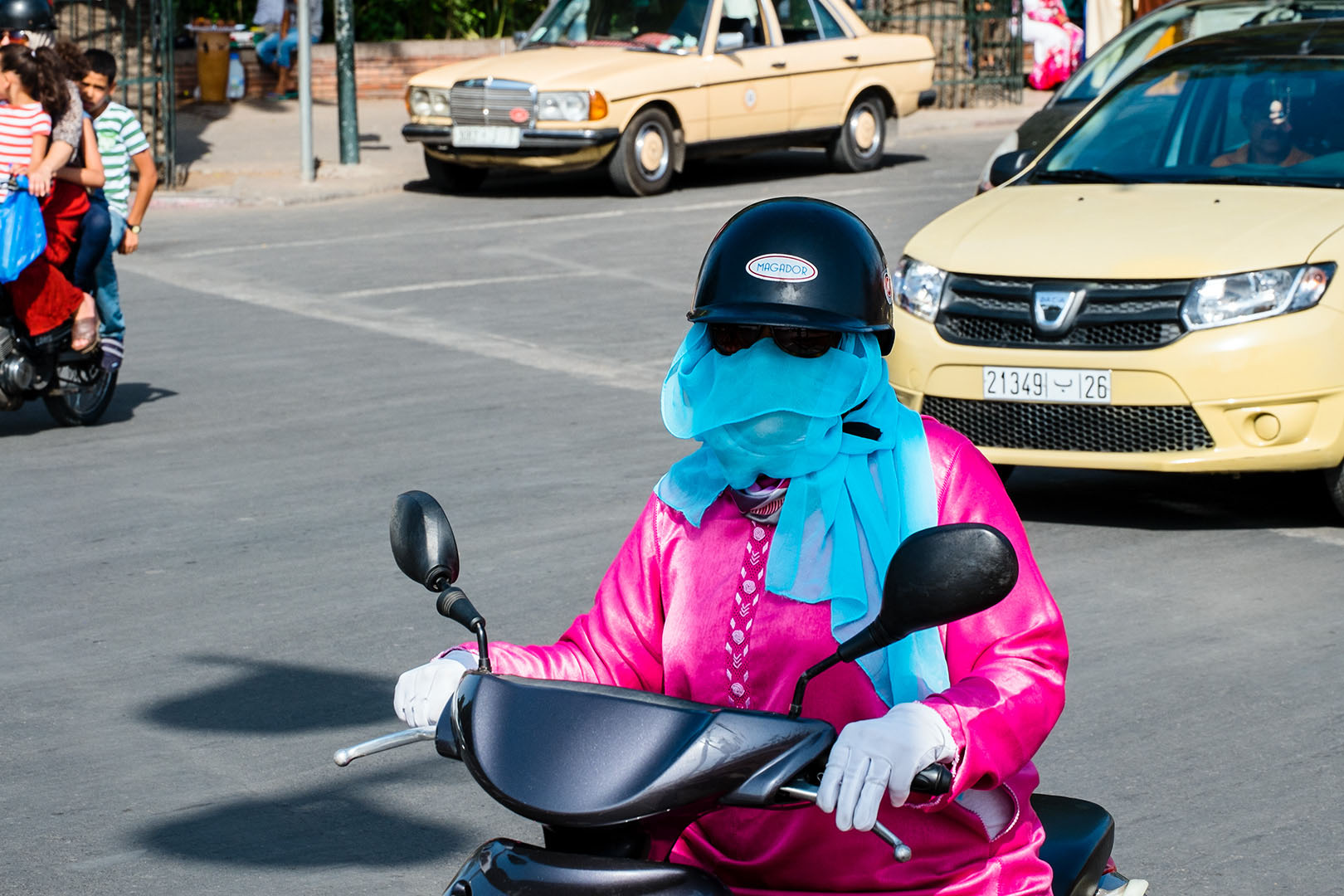Woman on Scooter in traffic with Pink tunic, blue headscarf, white gloves and dark glasses