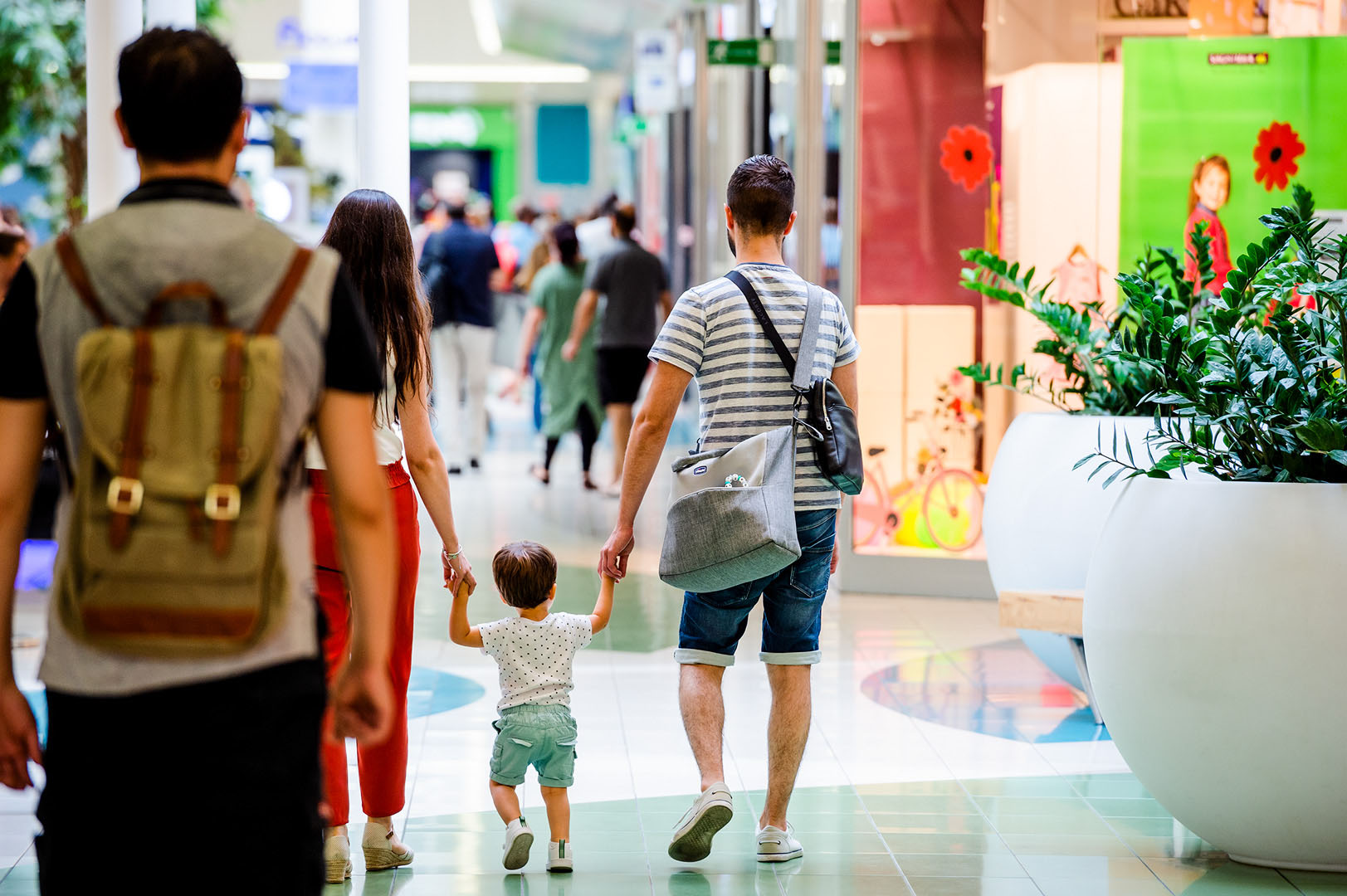 Mum & Dad holding hands with toddler shot from behind in busy shopping centre