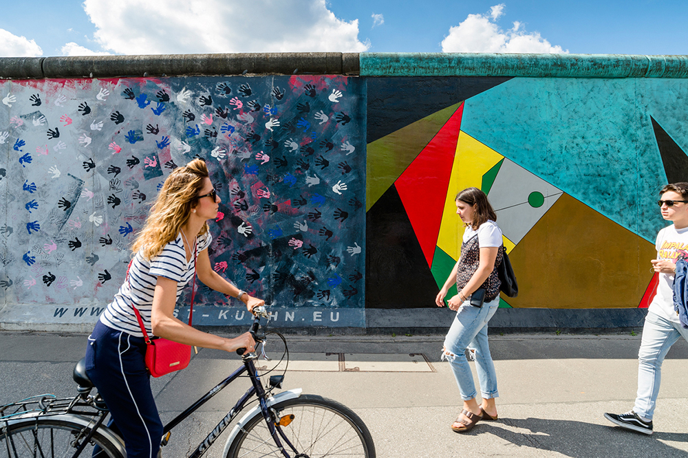 People walking & cycling in front of a section of the Berlin Wall at Eastside Gallery