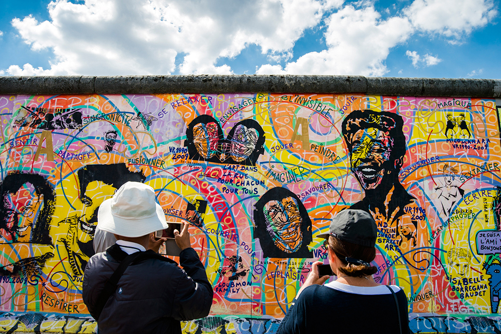 Two female tourists in hats from behind taking pictures of a section of the Berlin Wall at Eastside Gallery