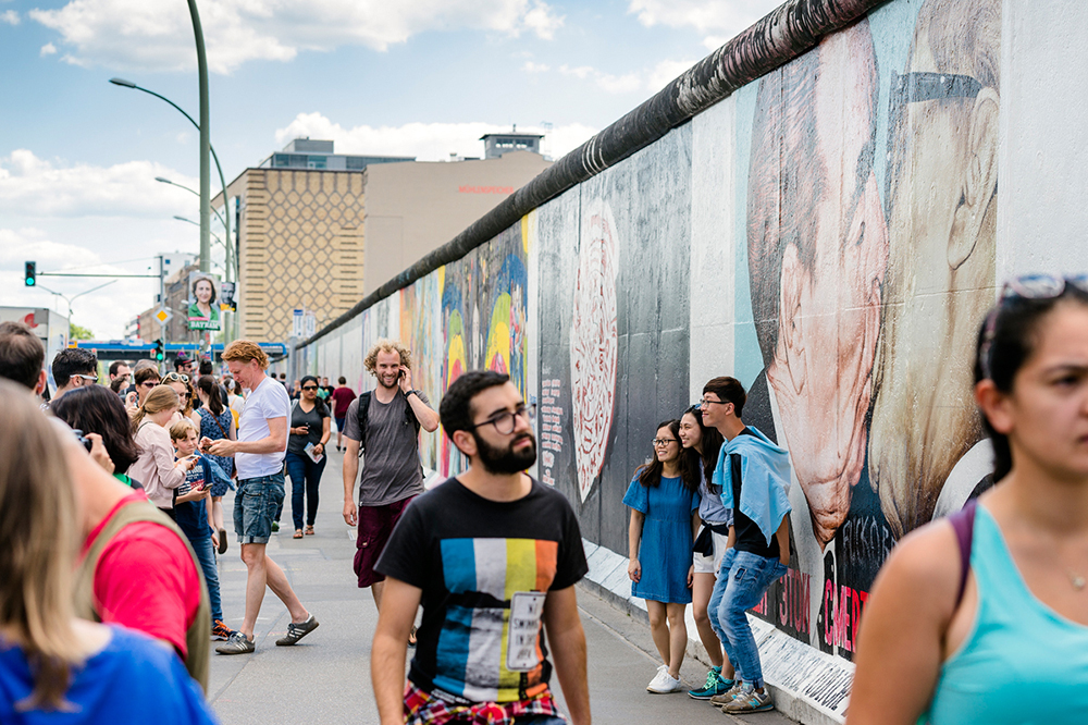 Three East Asian tourist friends being photographed next to a famous section of the Berlin wall surrounded by other tourists
