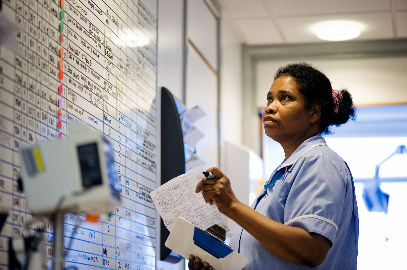 Black NHS nurse looking at patient information on whiteboard at nursing station