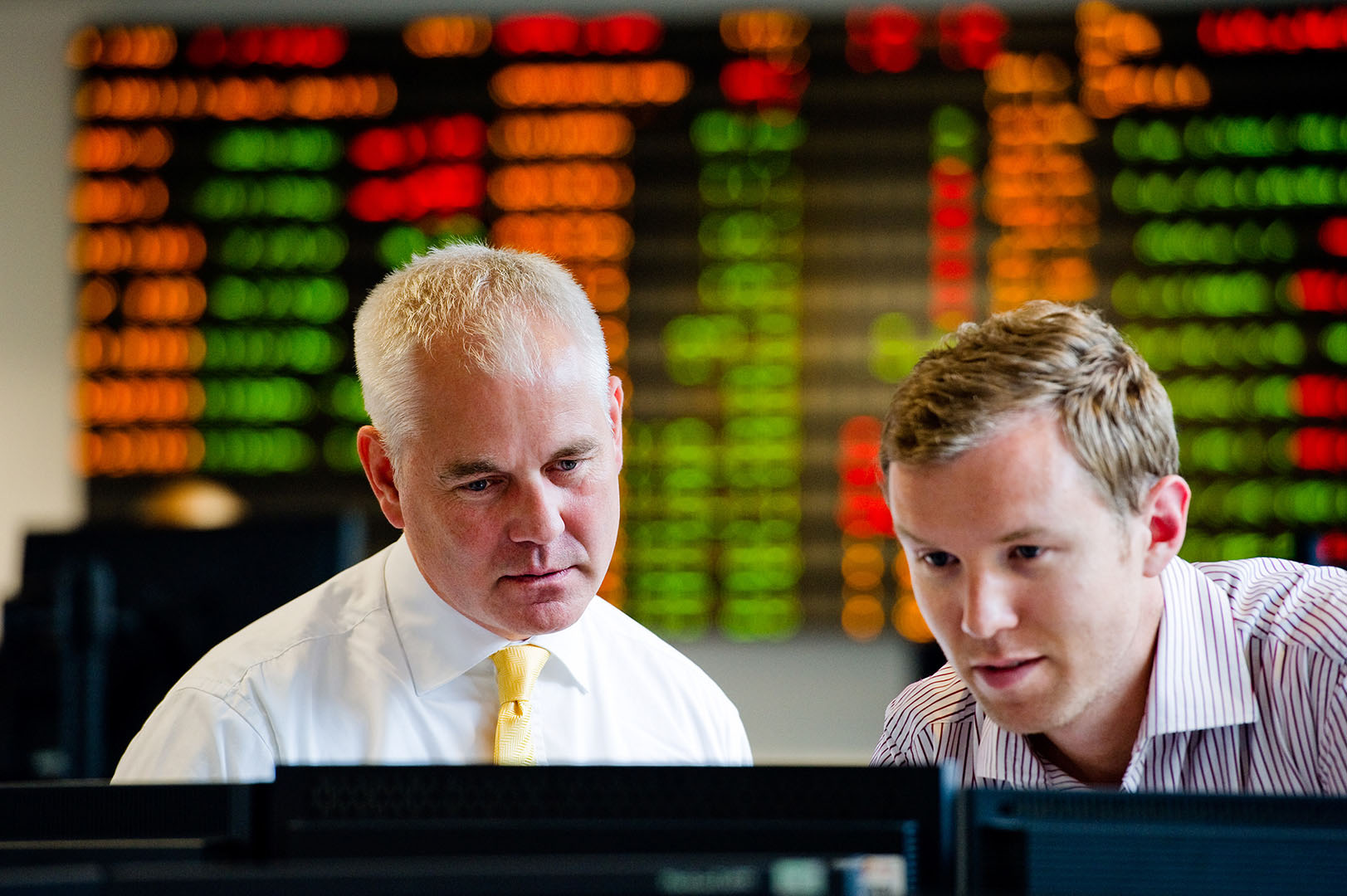 Two male colleagues looking at screen on trading floor with trading board in the background