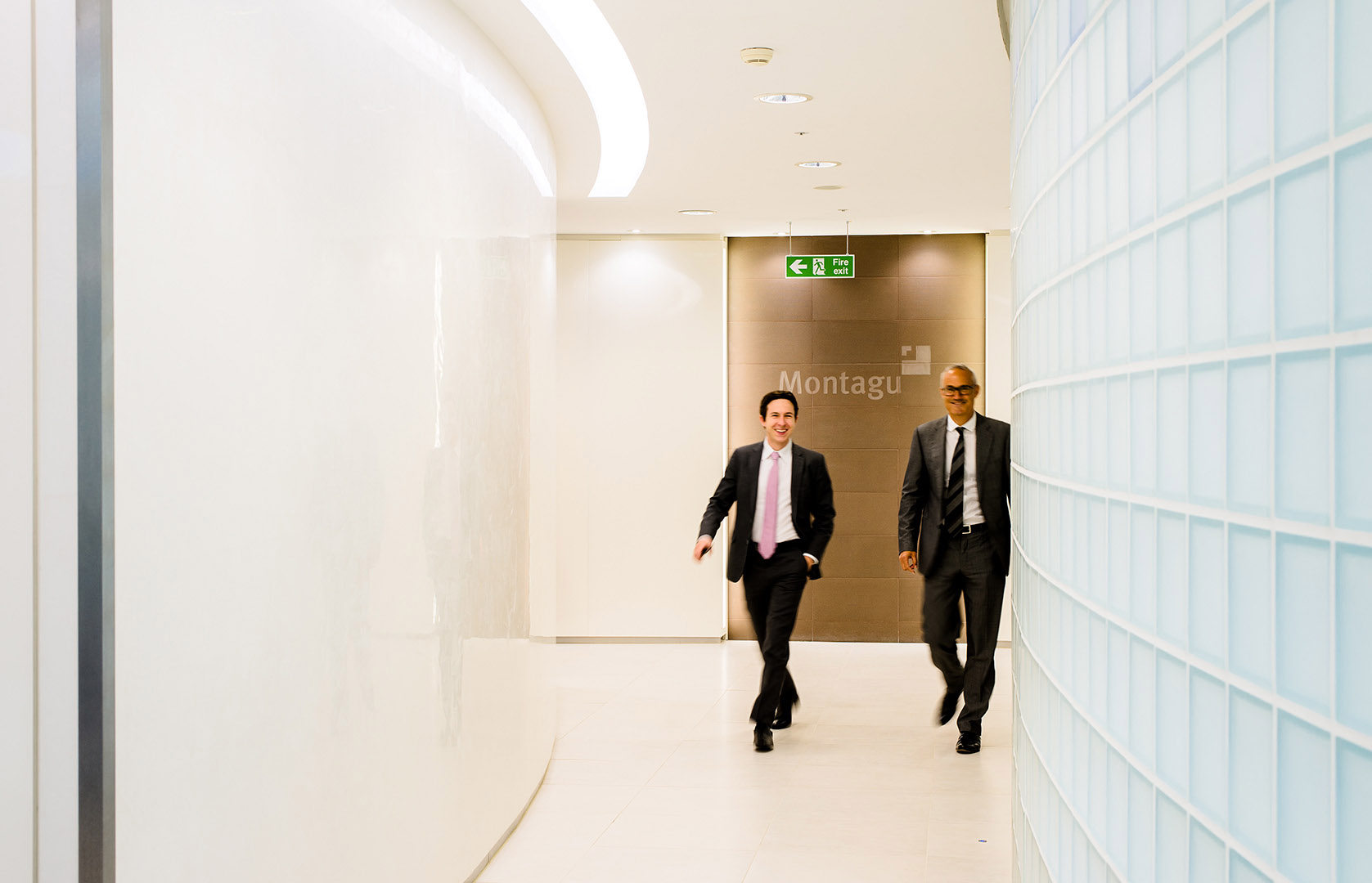Two businessmen walking towards camera in office corridor with glass bricks