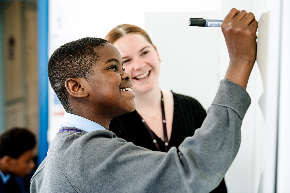 Young Black Male student writing on whiteboard with teacher in the background