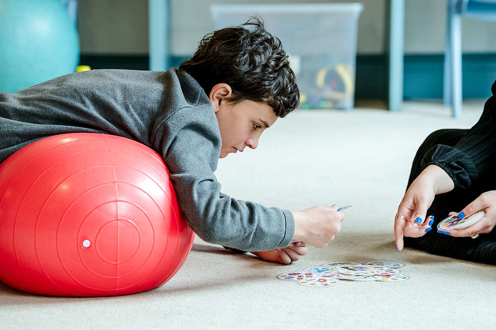 Young student playing card game lying on red exercise ball in therapy session